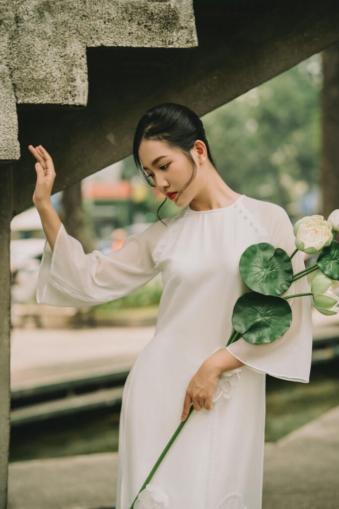 Graceful woman in white dress holding lilies under stairs, captured outdoors with a serene expression.